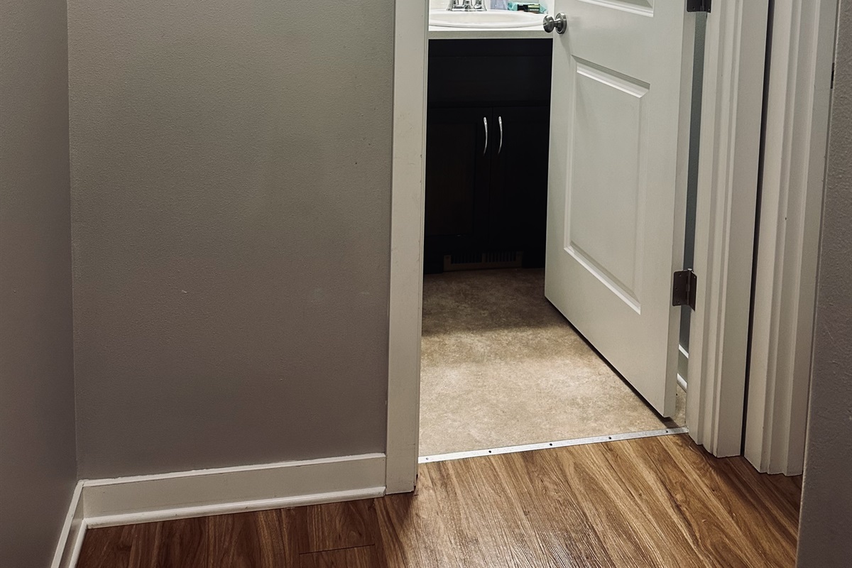A view down a small hallway featuring a partially open door leading to a bathroom with light-colored flooring, a dark cabinet beneath a sink visible inside, and brown wooden laminate flooring in the foreground.