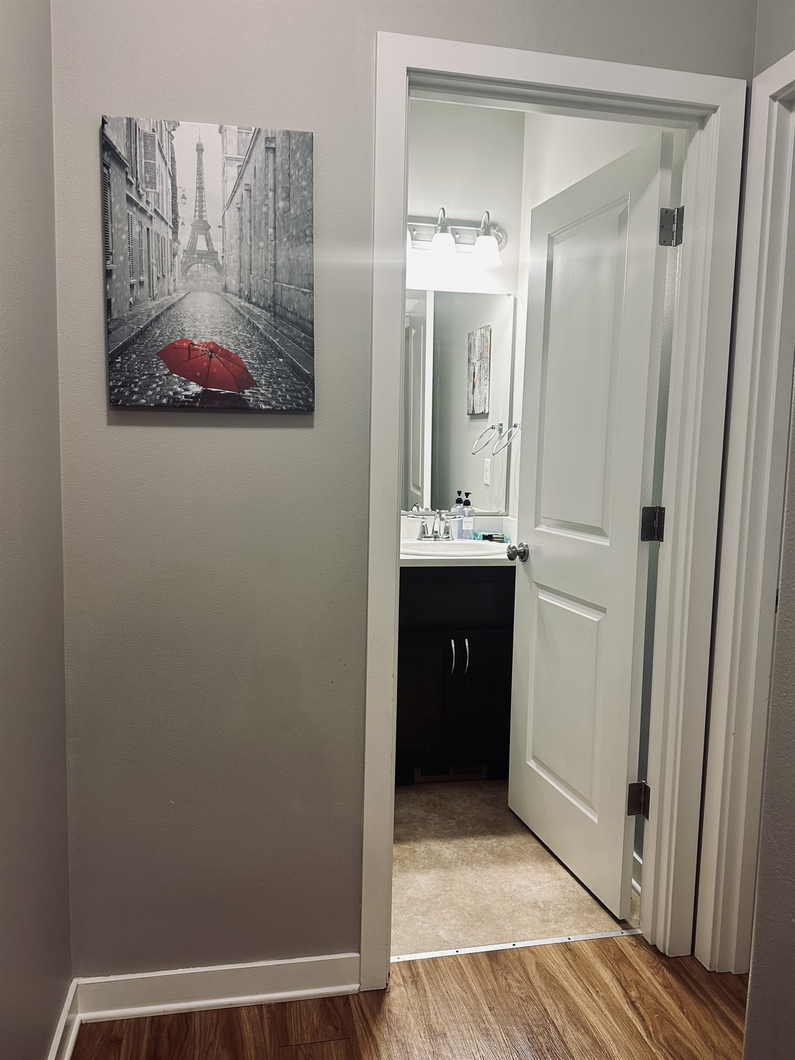 A bathroom doorway is visible, framed by white walls and doors, leading to a sink area with a flourescent light above and a mirror reflecting a black and white artwork featuring an Eiffel Tower and a red umbrella.