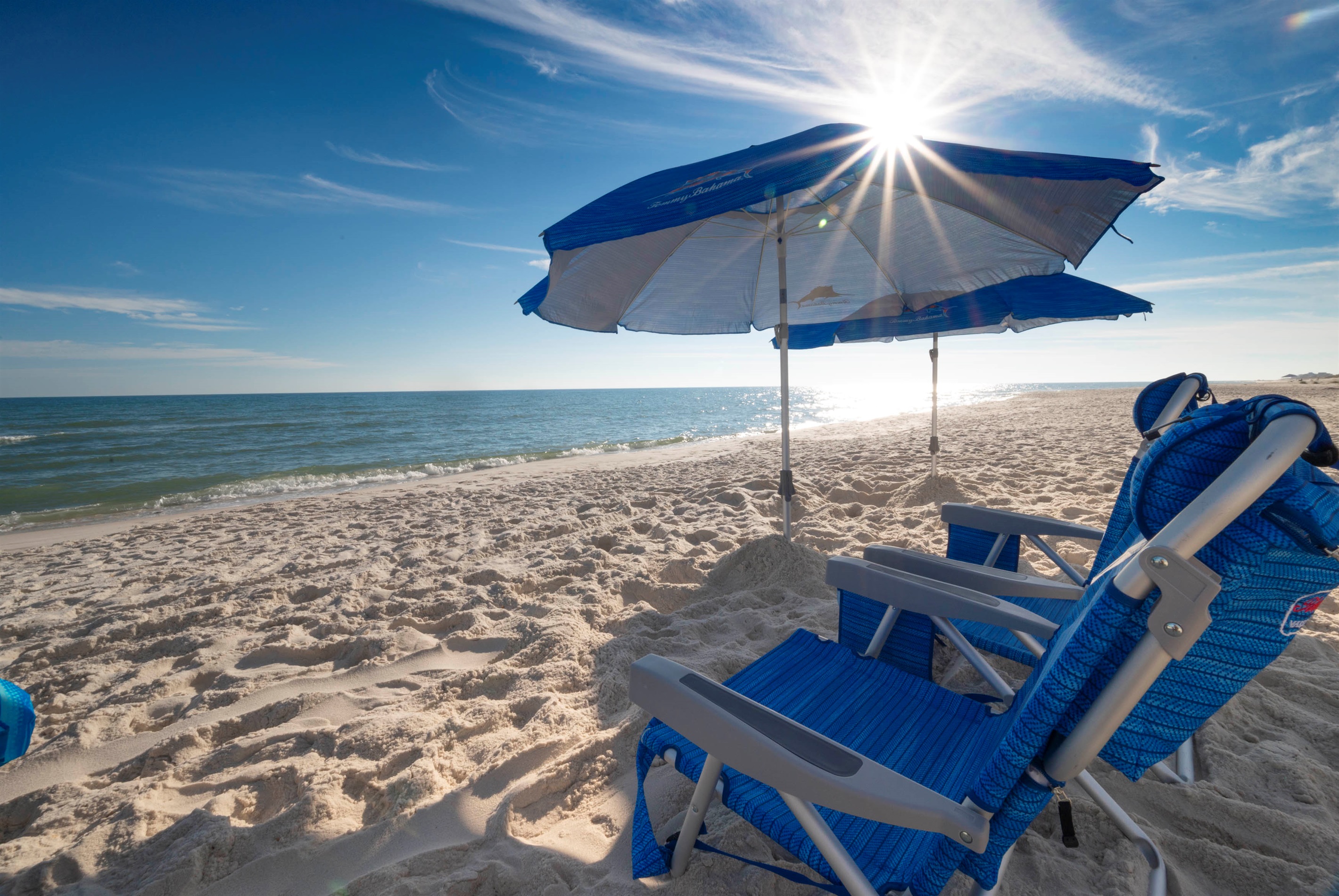 Relax in comfort with blue chairs and an umbrella, basking under sunny skies by calm waves on a sandy shore.
