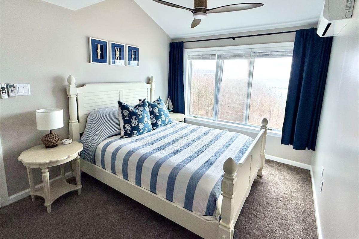 This coastal-themed bedroom features a white bed frame with blue-striped bedding and navy curtains. A bedside table, nautical wall art, and a ceiling fan complete the space with views of the outdoors.