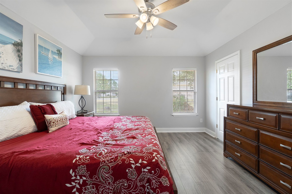 Unwind in this spacious master bedroom featuring a luxurious king bed, rich wood furniture, and elegant red bedding with beach-themed accents. Natural light streams through the large windows, creating a calm and inviting atmosphere for rest and relaxation.
