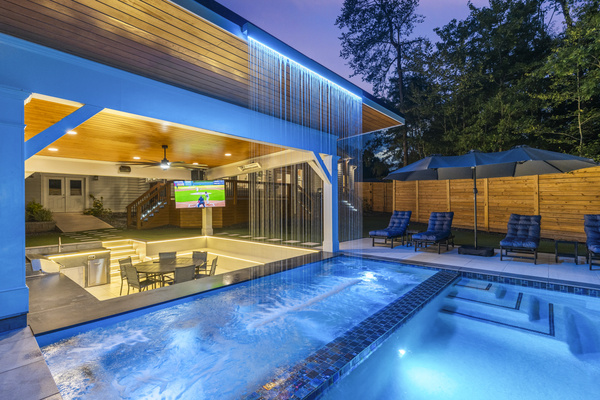 Luxury Lake Lanier living at Pool Villa Lanier: resort-style pool, cascading water feature, covered outdoor entertainment, dining, plush loungers, and a large-screen TV create the ultimate private retreat for relaxing and family bonding.
