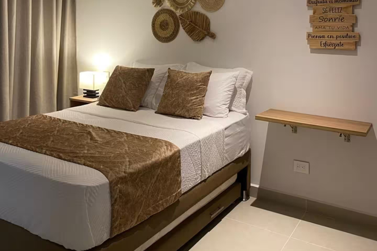 La ropa de cama en tonos neutros, la decoración de pared tejida y la suave iluminación junto a la cama crean un refugio tranquilo con un relajado ambiente de apartamento tipo resort.