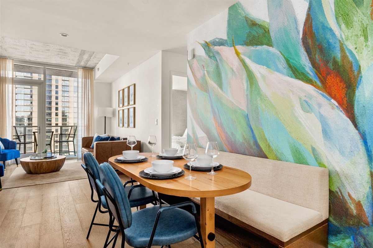 A chic dining area featuring a light wood oval table paired with a built-in bench and modern blue chairs. A vibrant, oversized floral mural serves as a striking backdrop, while the open floor plan offers a view into a bright living room and a balcony with