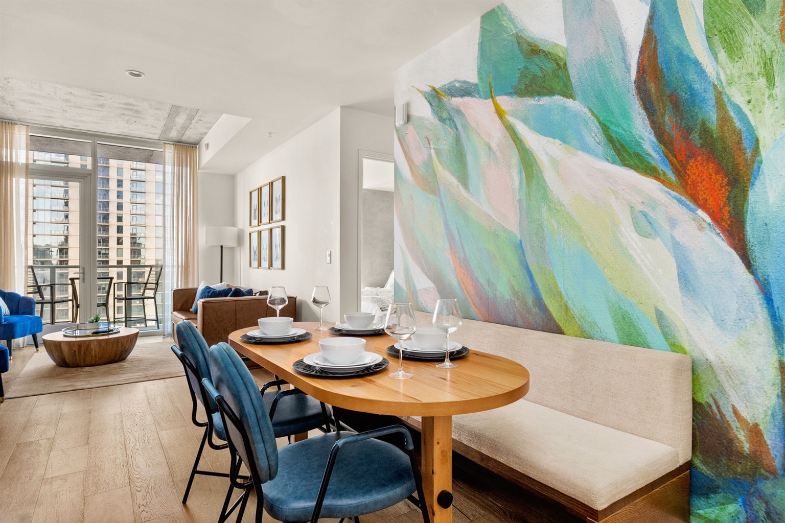A chic dining area featuring a light wood oval table paired with a built-in bench and modern blue chairs. A vibrant, oversized floral mural serves as a striking backdrop, while the open floor plan offers a view into a bright living room and a balcony with