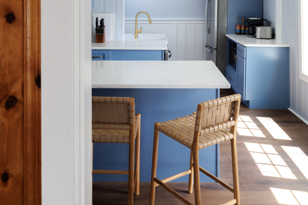 Blue-gray island and rattan stools make the kitchen a natural gathering spot even before the meal — someone always ends up perched here all evening.
