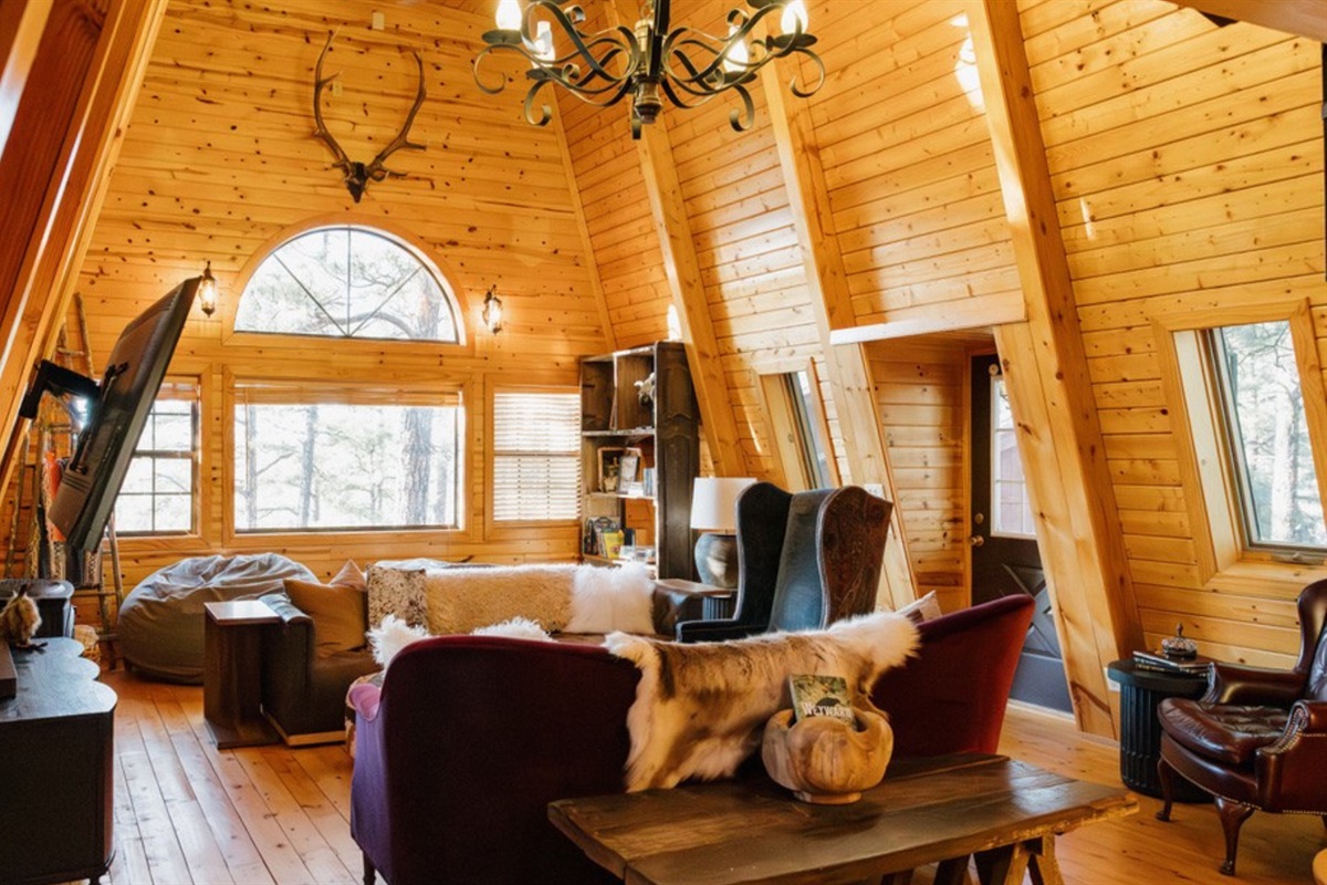 Spacious A-frame living room with dramatic vaulted ceilings, statement chandelier, and multiple seating areas—ideal for gathering, movie nights, and relaxing in a warm, rustic White Mountains cabin retreat.