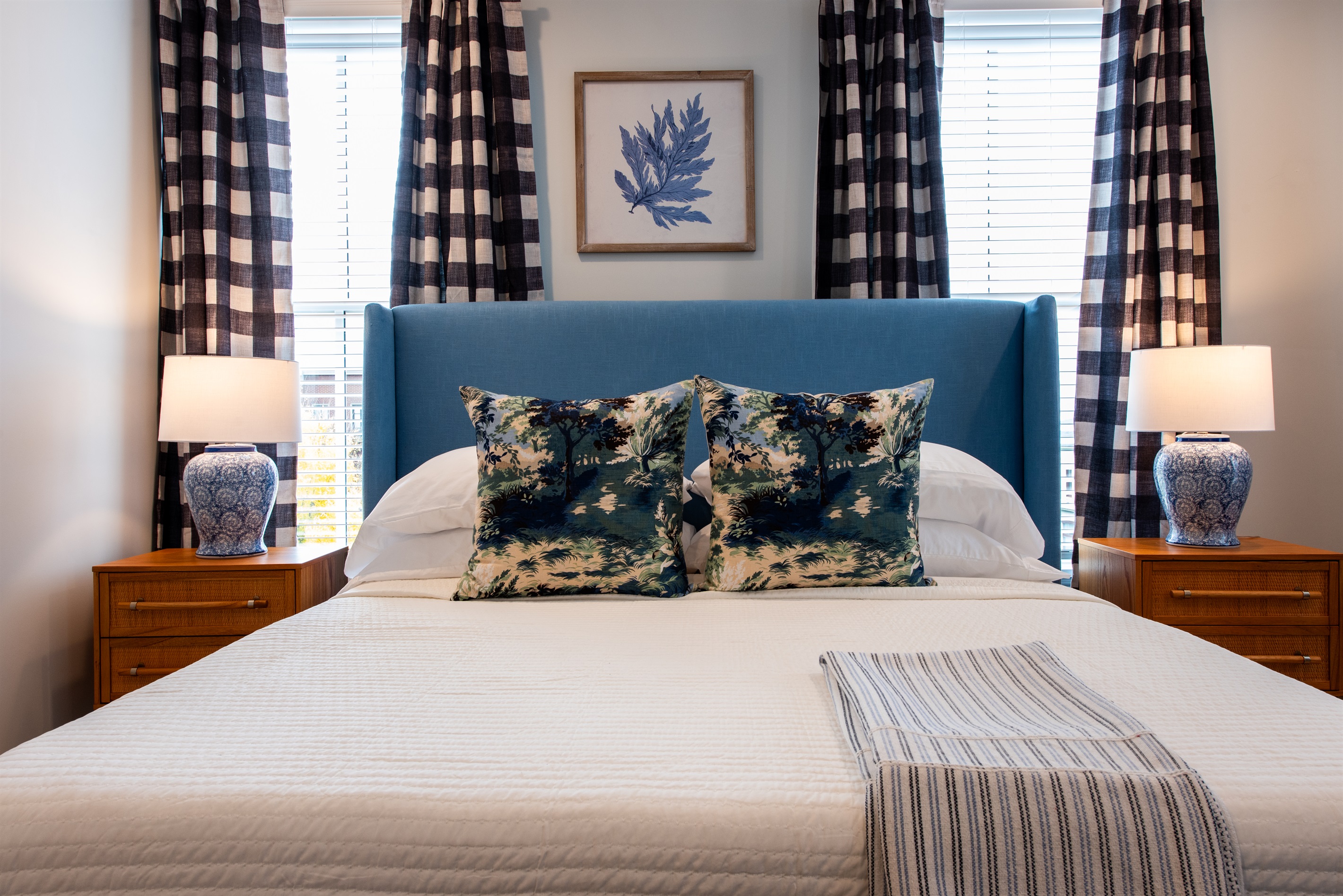 Unwind in this tastefully decorated bedroom featuring a plush blue headboard, elegant decor, and cozy furnishings. The room's chic design with checkered curtains and botanical art creates a perfect haven for your stay on the Bourbon Trail.