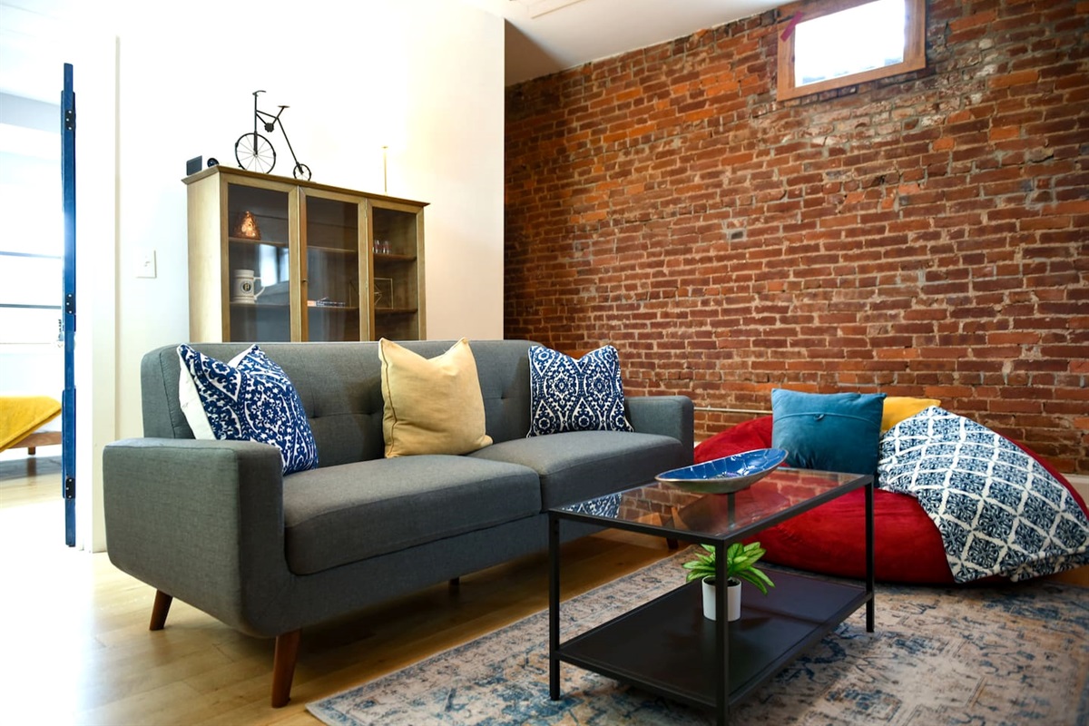 This living room features a projector screen, comfy couch, and a chic brick wall—plus a cozy rug and bean bag for the perfect movie night or chill session.
