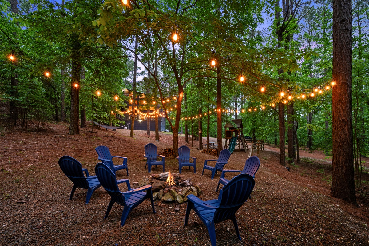 As dusk settles in, the fire pit area becomes a cozy gathering spot, with string lights overhead adding a warm and inviting glow.
