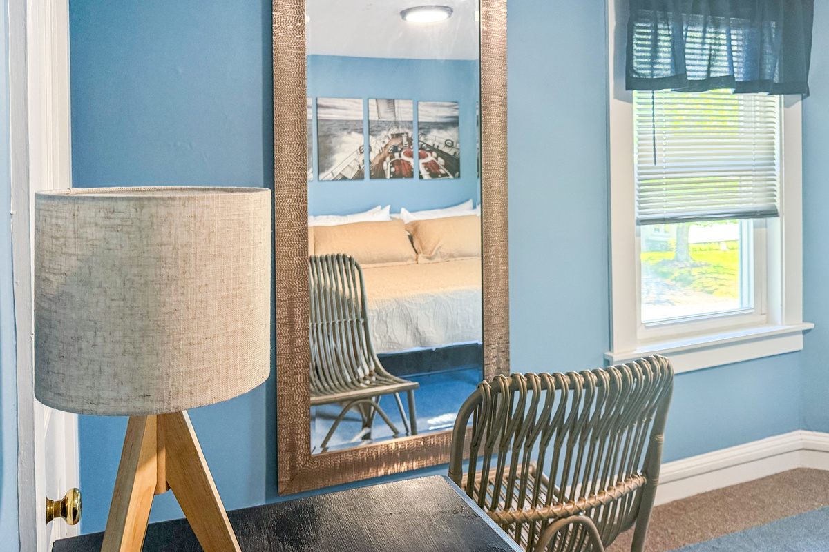 The primary bedroom features this full-length mirror and woven rattan chair, providing a dedicated space for getting ready or applying makeup. The reflection reveals the maritime artwork and memory foam king bed that define the rest of the room.
