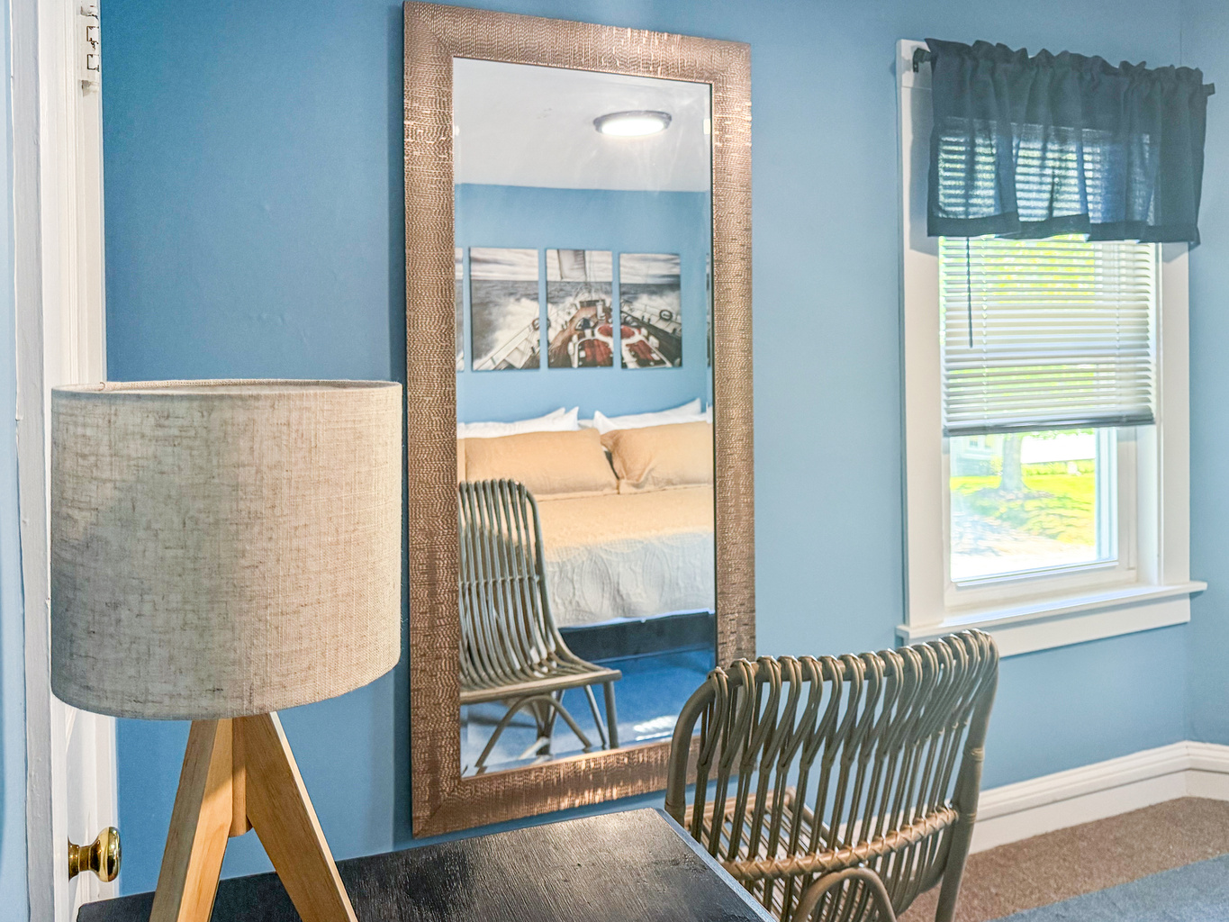 The primary bedroom features this full-length mirror and woven rattan chair, providing a dedicated space for getting ready or applying makeup. The reflection reveals the maritime artwork and memory foam king bed that define the rest of the room.