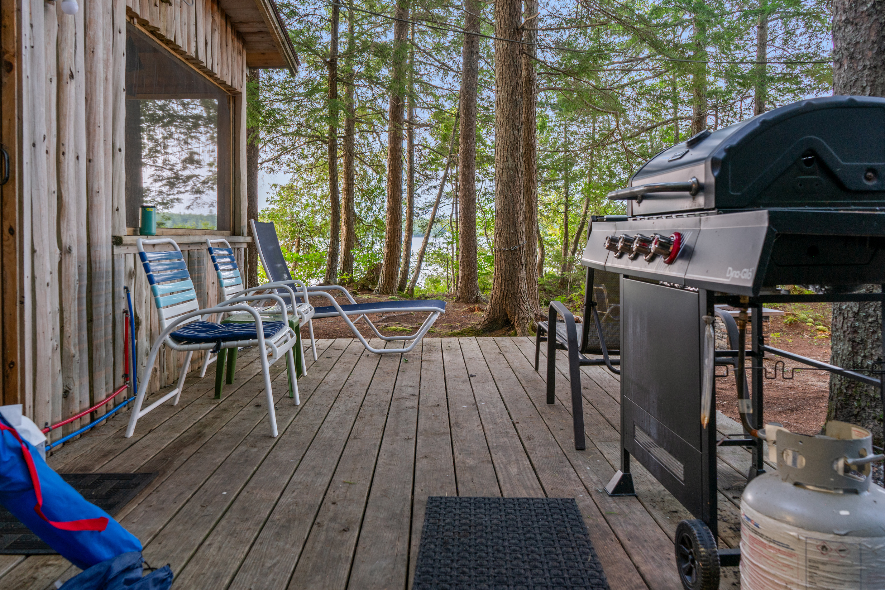 Close-up of the kitchen deck at Loonrise Point! Features a Dyna-Glo propane gas grill with tank, patio seating, and water views through the trees. Weathered deck boards and exposed plumbing on historic cedar siding on Beech Hill Pond in Otis, ME.