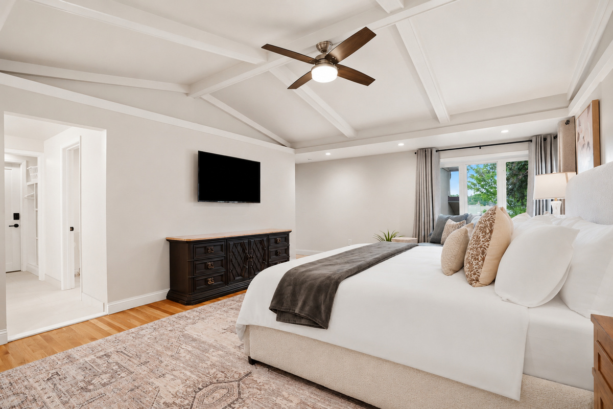 Relax in this bright and spacious bedroom, featuring vaulted ceilings, a ceiling fan, large windows, and elegant decor for ultimate comfort and rest.
