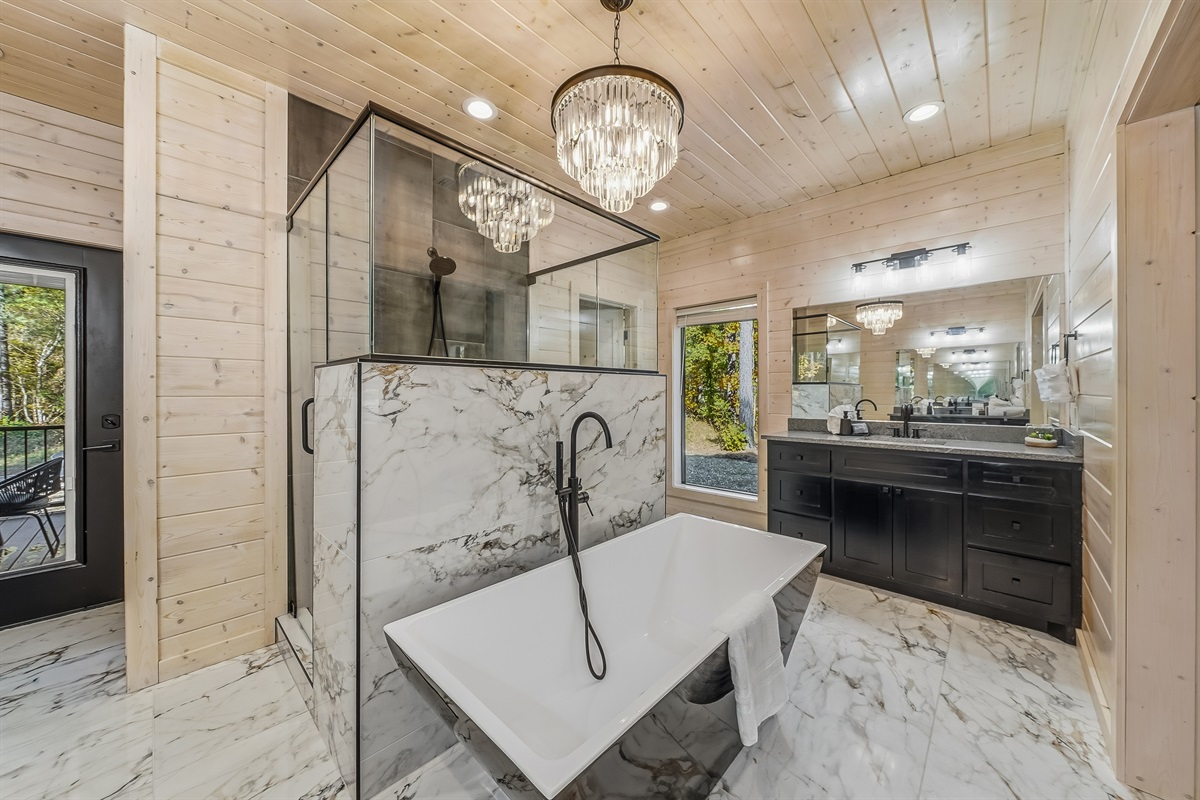 A luxury bathroom centerpiece featuring a sculptural soaking tub under a crystal chandelier and surrounded by forest framed windows.