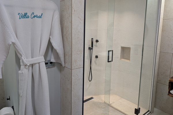 Premium custom-embroidered Villa Coral bath robe hanging next to a glass steam shower enclosure with digital touch control panel in the primary suite bathroom in Dorado Puerto Rico.
