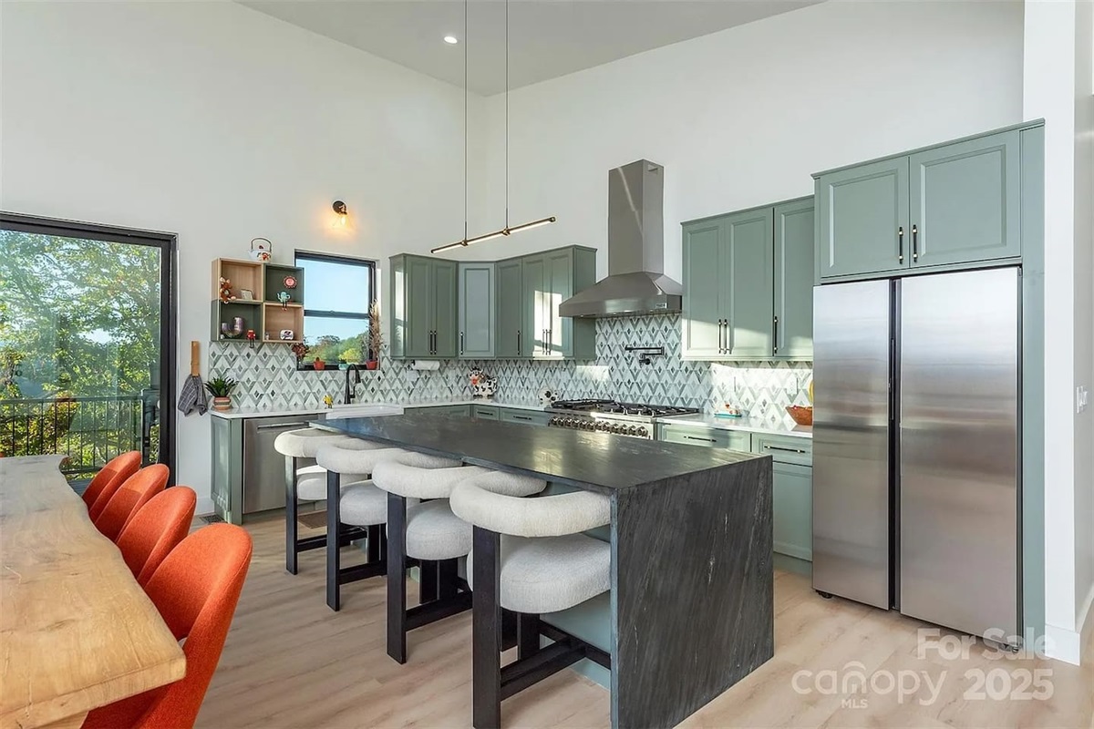 Modern, fully equipped kitchen featuring custom sage-green cabinetry, stainless steel appliances, a spacious island with bar seating, and stylish tile backsplash, ideal for cooking, entertaining, and gathering during your stay.