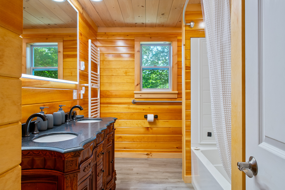 Refresh in the gorgeous primary bathroom featuring a large double vanity with an ornate wood base and dark stone countertop. The space is detailed with an illuminated LED mirror, beautiful log-cabin wood walls, and a full bathtub/shower combo.