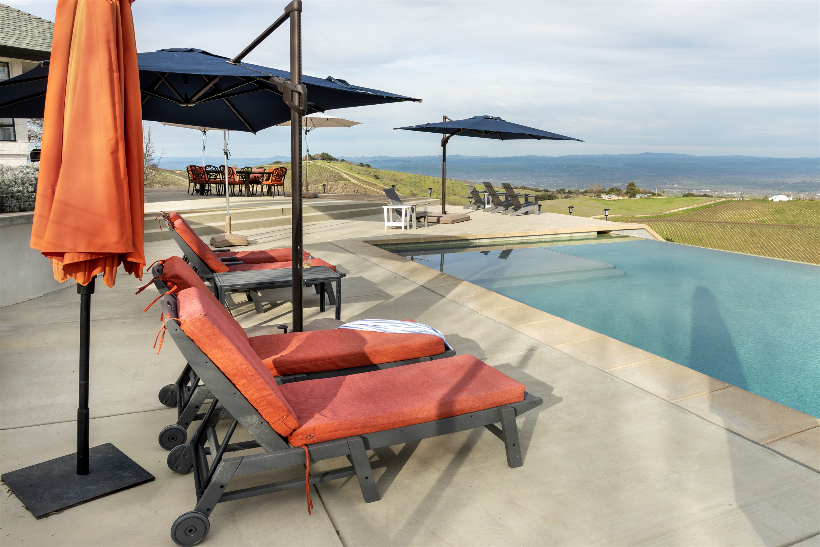 The full picture — cushioned loungers by the pool, the outdoor dining table beyond, Adirondack chairs at the infinity edge, and miles of wine country in every direction. Everything you need, nothing you don't.