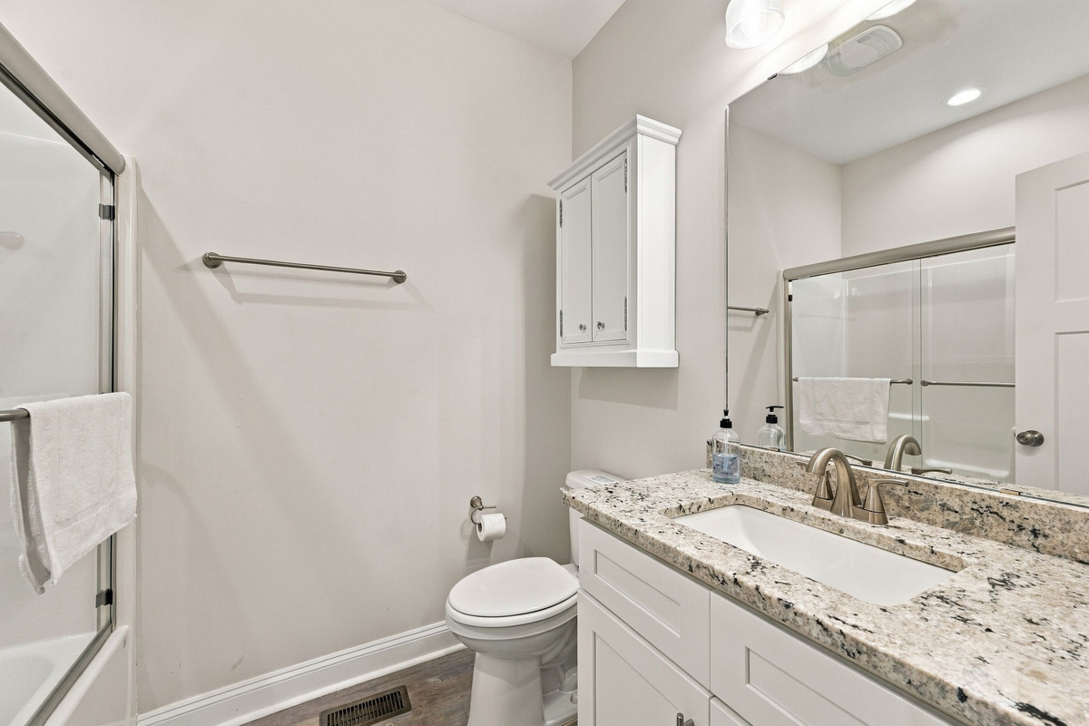 Full bathroom featuring a white single-sink vanity with a granite countertop, large vanity mirror, wall-mounted medicine cabinet, toilet, and a sliding glass door shower enclosure.   