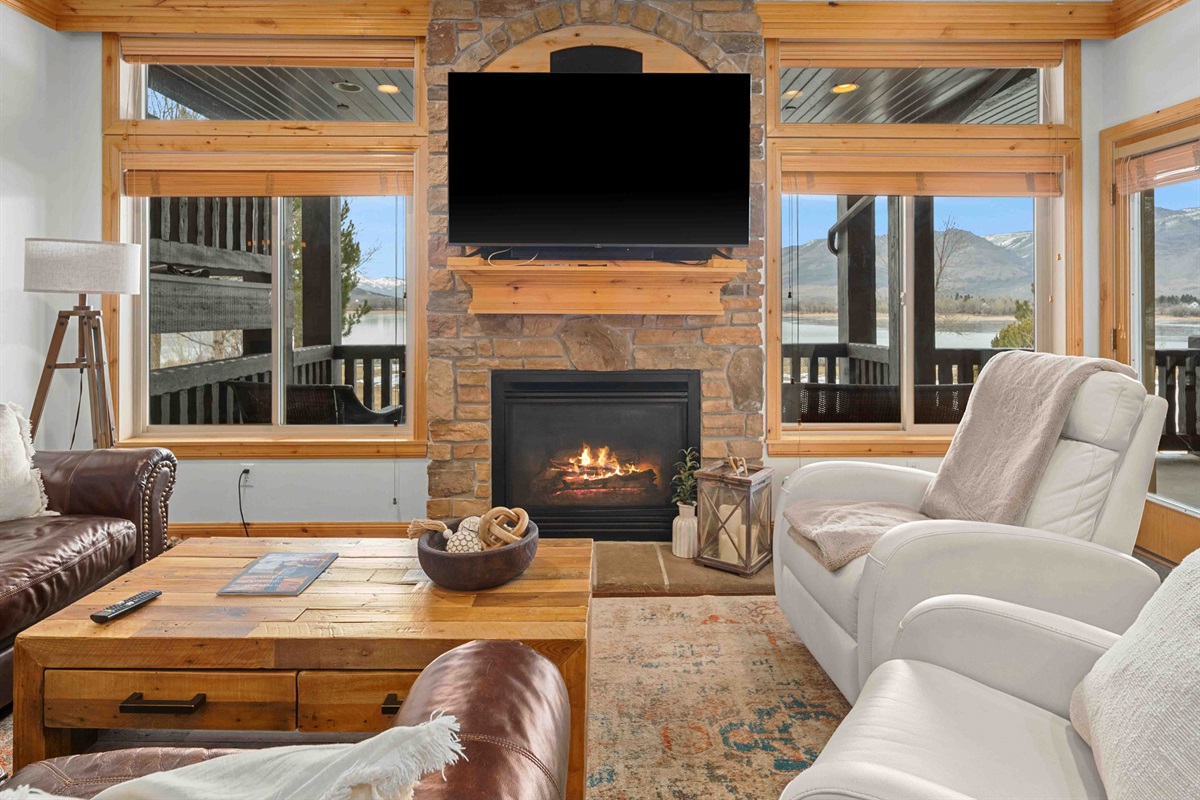 Cozy living room featuring a stone fireplace, large windows, and warm natural light.