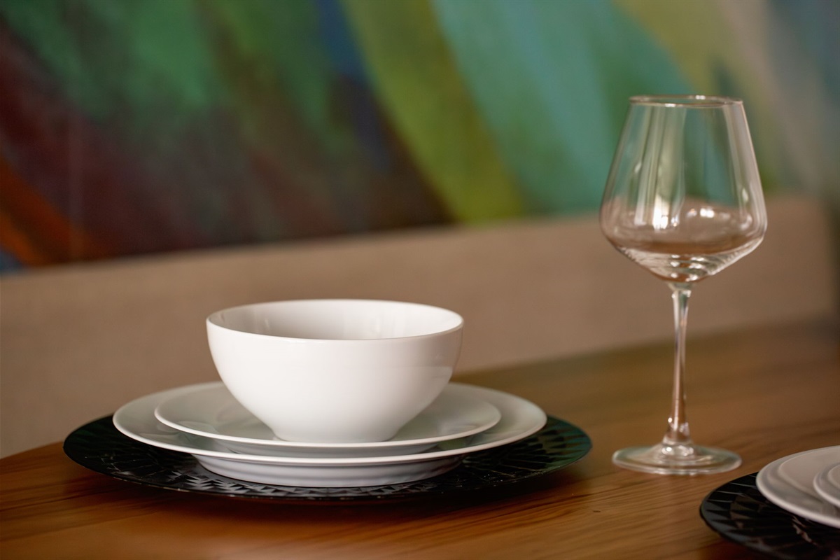 A detailed shot of a dining table setting, showcasing a stack of white dinnerware and a clear wine glass on a wooden surface, with a colorful, abstract painting visible in the soft-focus background.