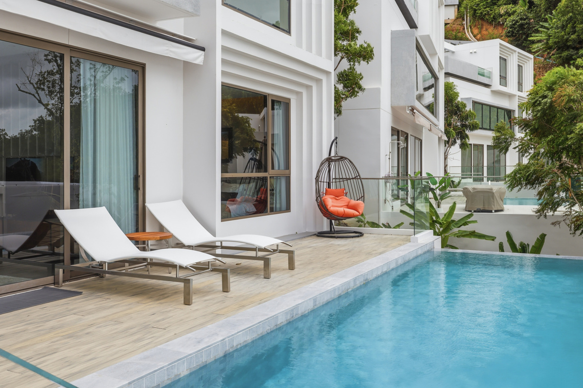 Pool deck with lounge chairs, hanging swing, modern white building, and access to interior spaces.