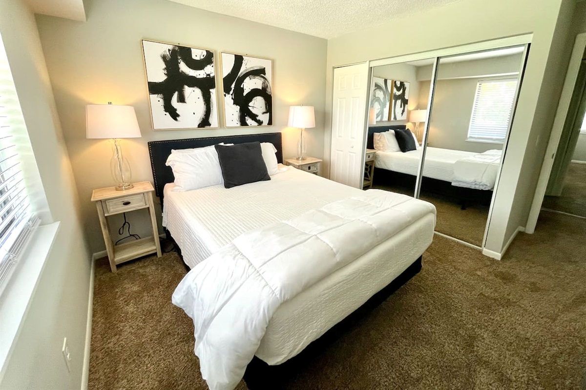 Stylish and serene bedroom with plush carpeting, a comfy queen bed, and mirrored closet doors. Thoughtful touches like matching nightstands, modern lamps, and bold black-and-white art make this a restful, inviting space for your short-term stay.