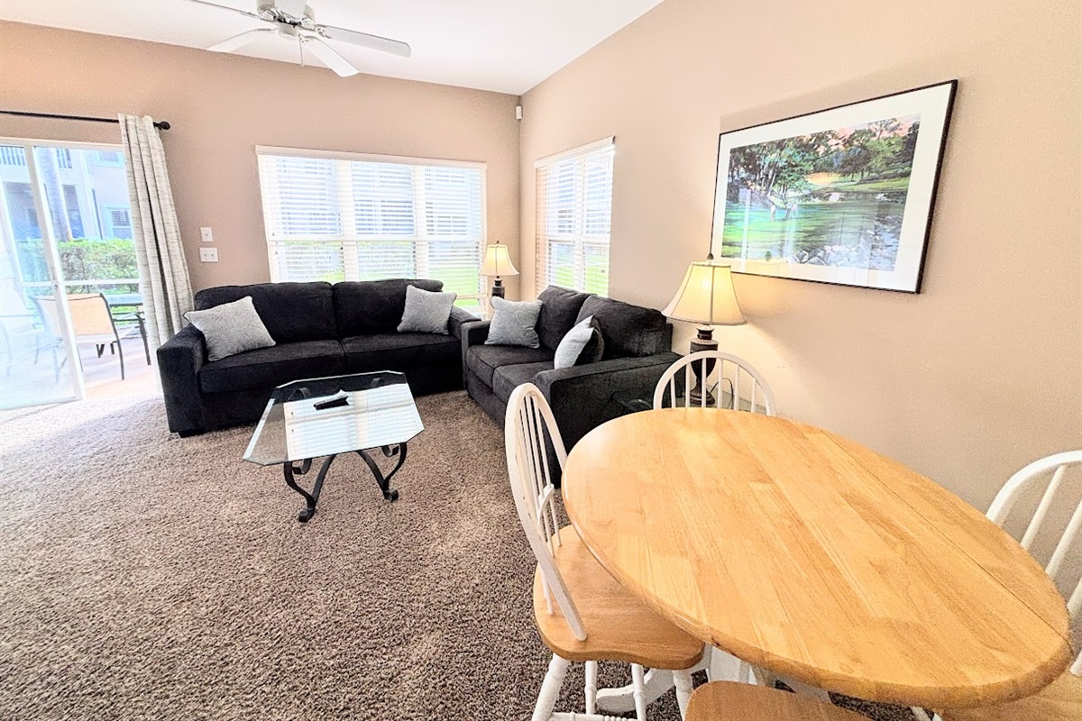 Spacious living and dining area with cozy seating, ceiling fan, round dining table, and natural light from the patio doors.