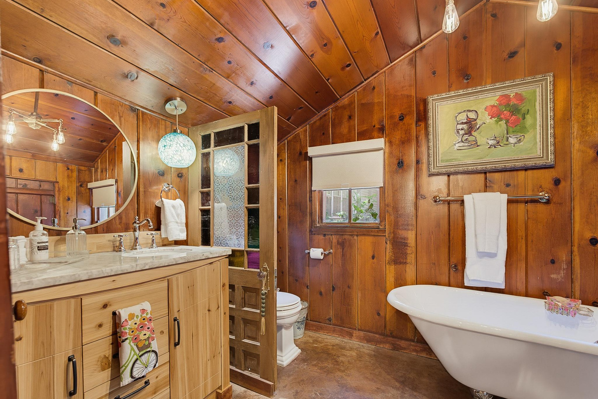 Papaw's Cabin spa-style bathroom with marble vanity, round mirror, stained-glass door, and vintage clawfoot tub. Rustic knotty pine charm meets modern comfort in Waco, Texas.