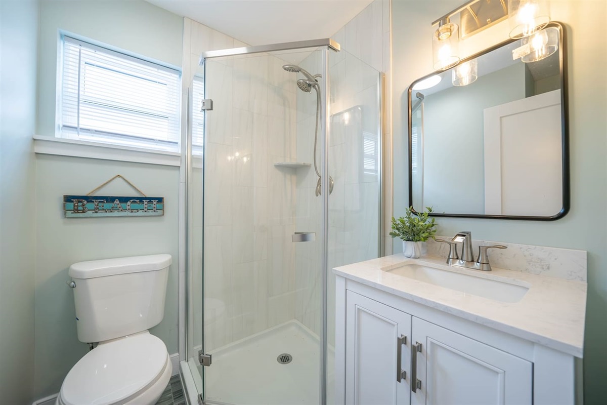 Fresh, light filled bathroom with a glass walk in shower, stone vanity, and bright lighting. Clean finishes and thoughtful layout create a comfortable space to refresh after the beach or get ready for the day.