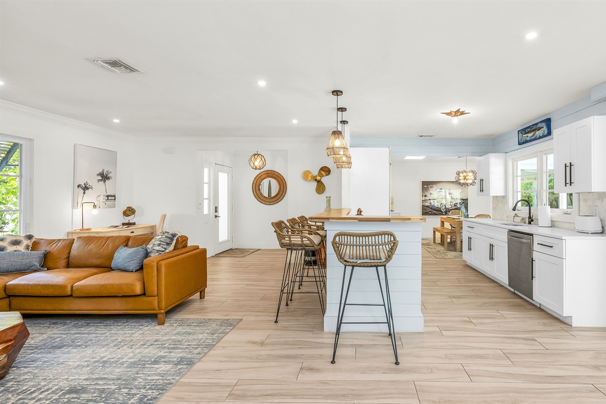 The bright and airy living room features a modern leather sectional, warm wood accents, and lush greenery views through large picture windows. Stylish touches like the surfboard décor and geometric coffee table create a relaxed coastal vibe.
