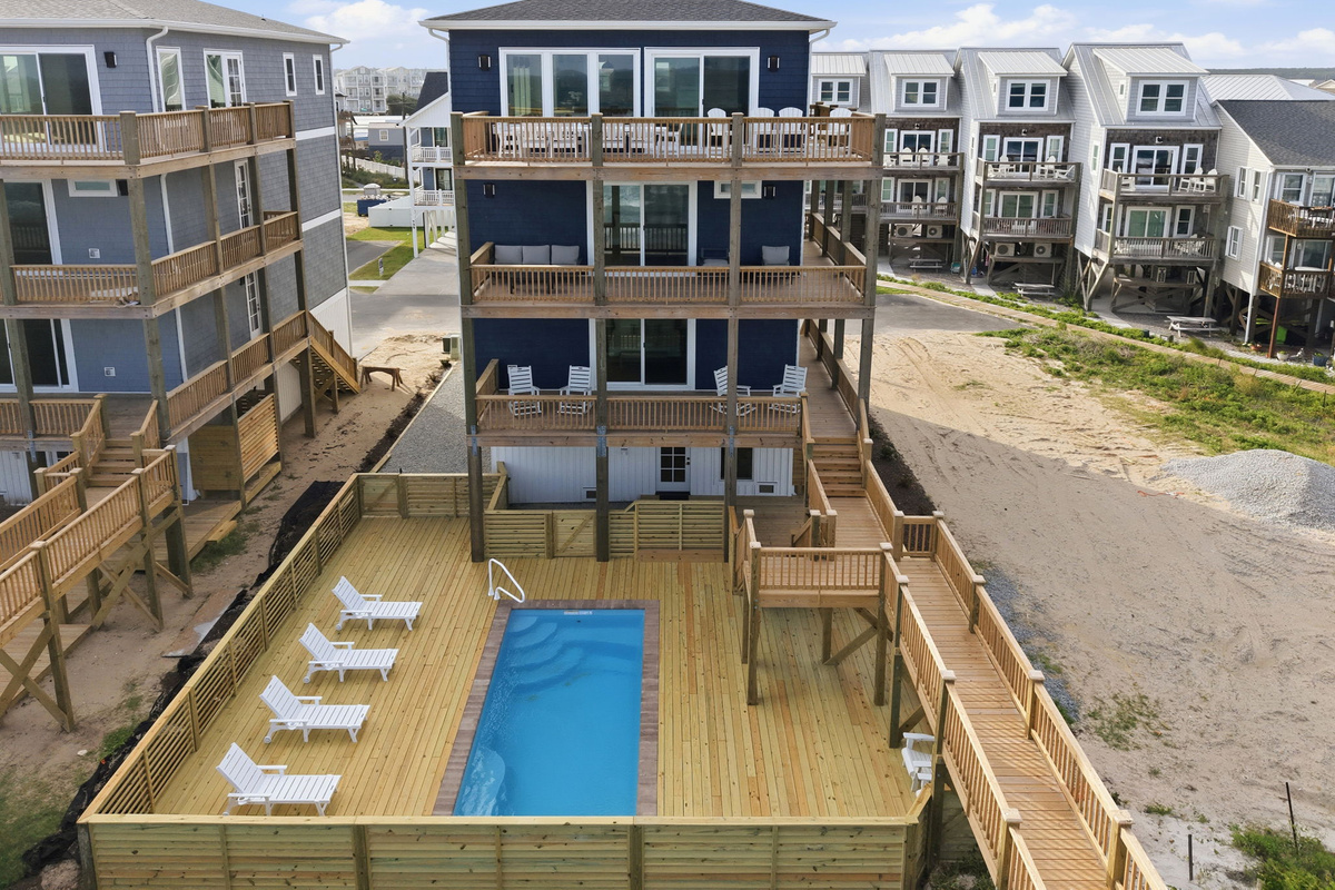 Aerial exterior view highlighting the pool deck area surrounded by multiple coastal homes, with lounge chairs, fencing, and elevated beachside walkways connecting the properties