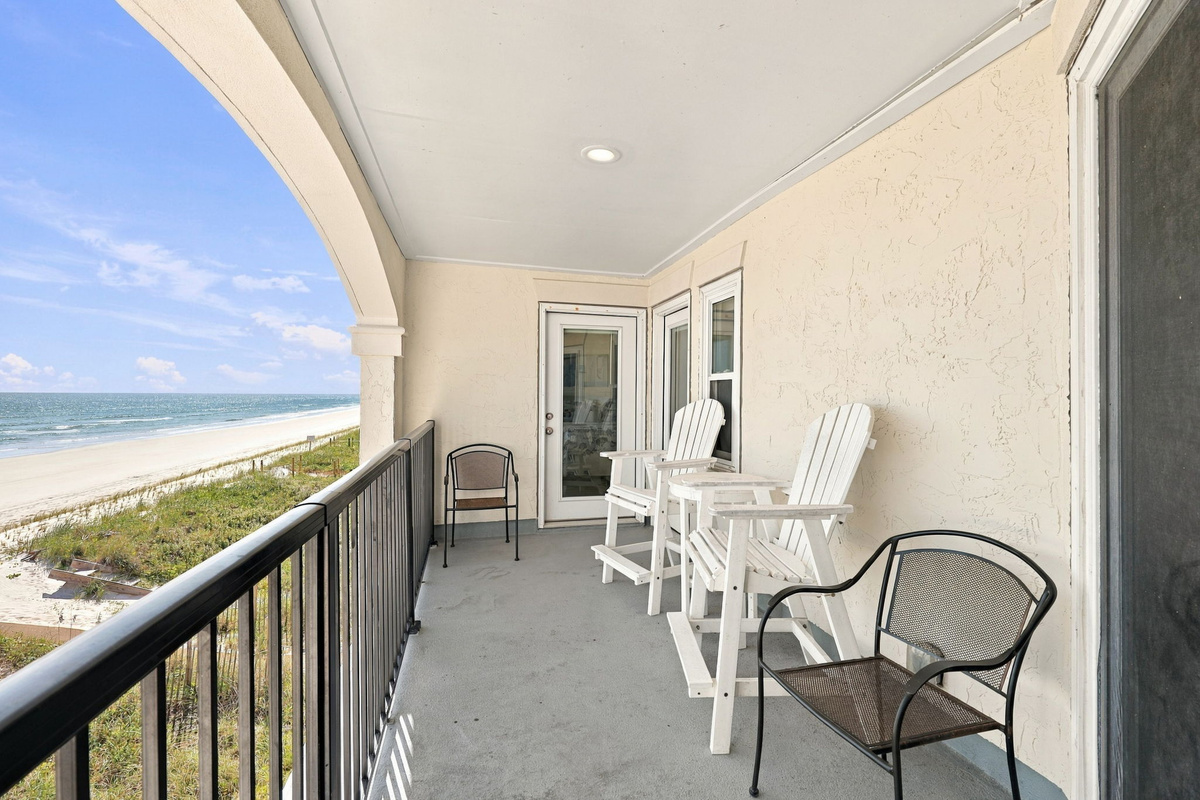 Covered balcony area lined with white tall Adirondack chairs and black metal seating, bordered by a dark railing overlooking the beach and ocean-horizon vistas.   