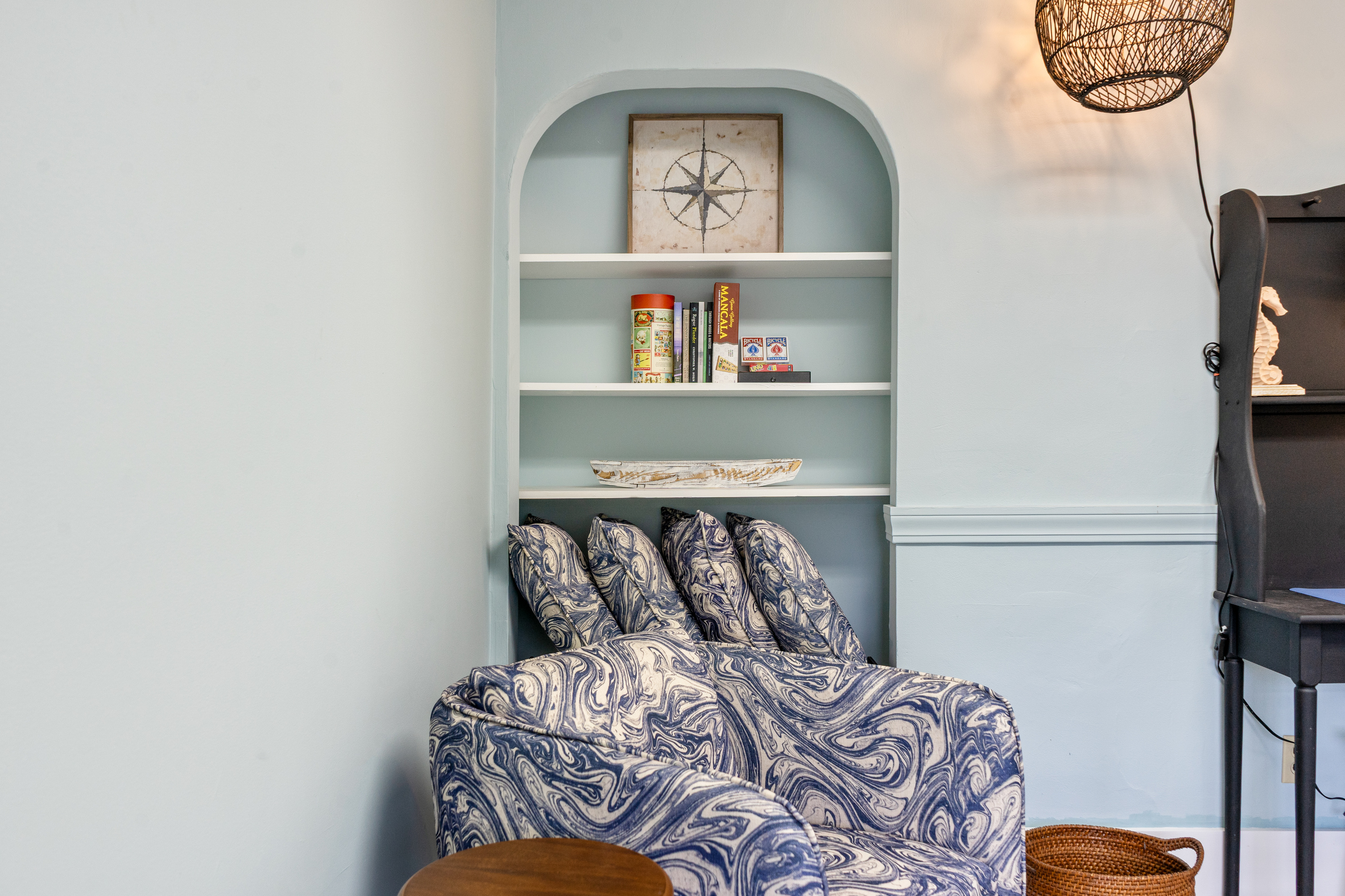 The compass den includes an arched built-in shelving alcove stocked with books and board games. Positioned above the patterned tub chair, the shelves display the room's namesake compass rose artwork along with classic family entertainment options.