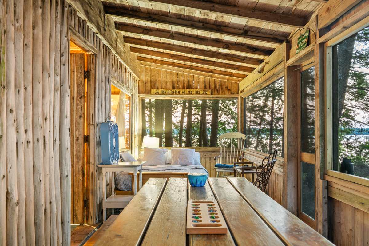 Full-length view of the screened porch at Loonrise Point! Wooden picnic table with board games flows down to the futon bed area overlooking Beech Hill Pond in Otis, ME. Rustic log-slab wall detail & open rafters create ideal porch living!