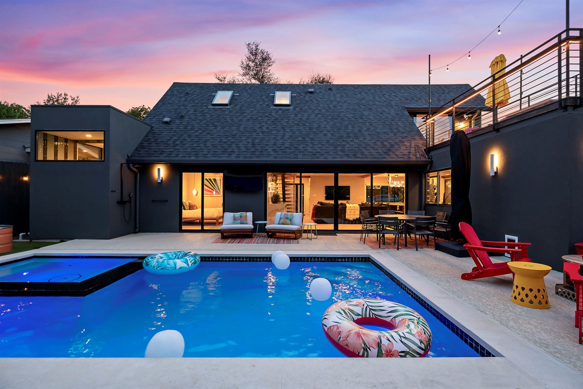 A luxurious backyard with a sparkling blue swimming pool, an adjacent hot tub, and a multi-level deck for dining and lounging, all set against a modern dark gray home with large glass doors and an impressive rooftop balcony.