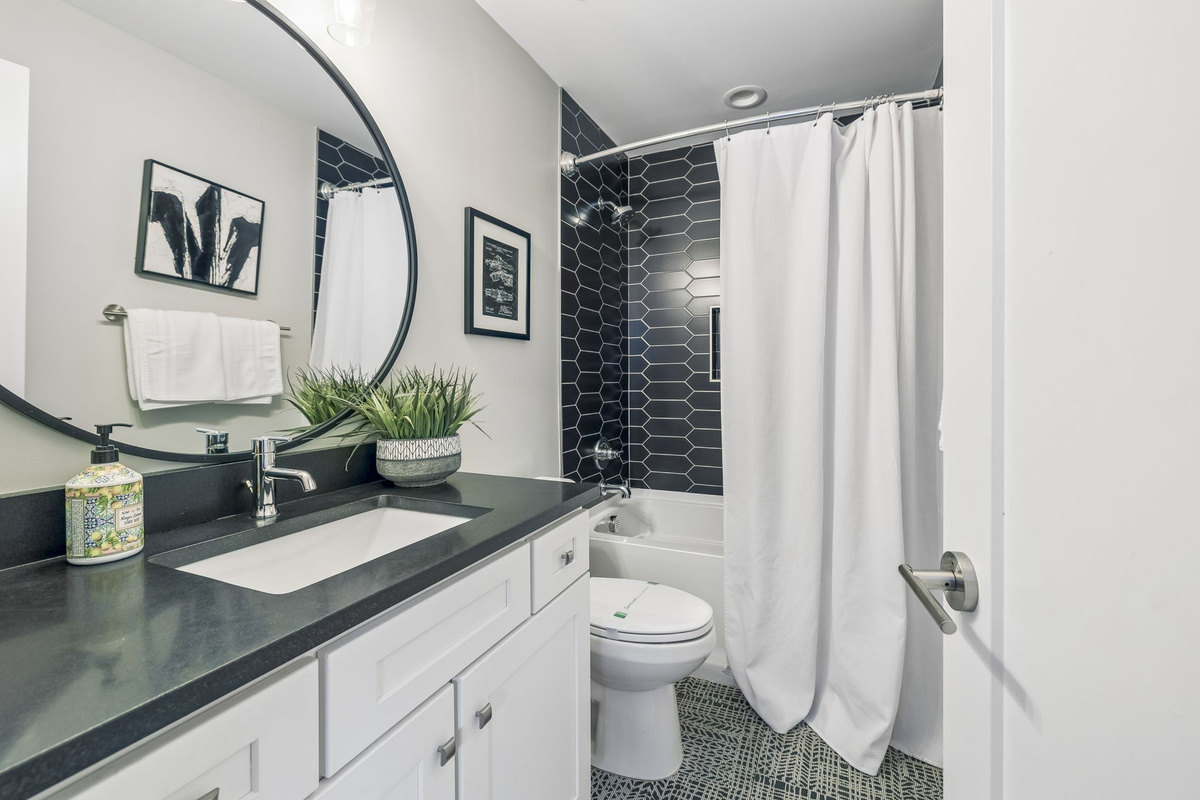 Striking black tile shower with geometric pattern and built in niche for essentials - Round mirror and dark vanity top add contrast and modern flair to the space - Bright lighting, framed art, and greenery finish the spa like look with style