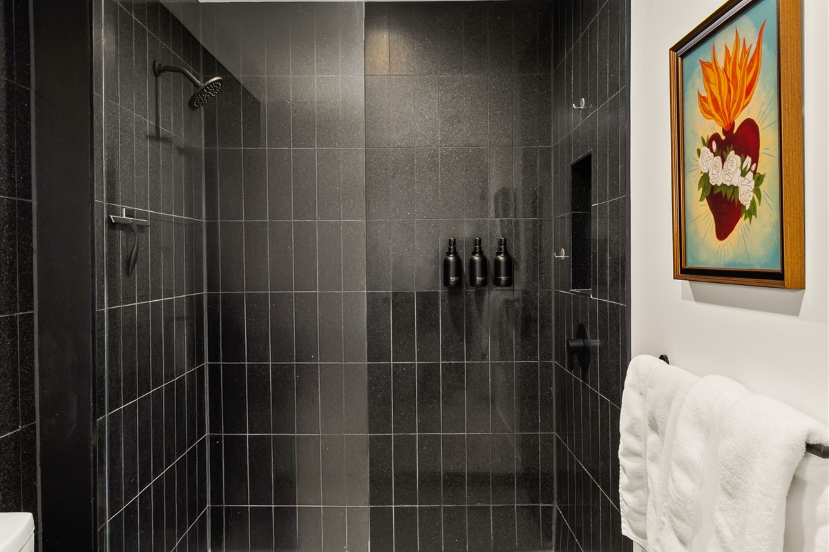 A spacious walk-in shower with sleek black tile, a built-in niche for toiletries, and a modern showerhead, complemented by an artistic, colorful painting of a sacred heart on the wall outside the shower.