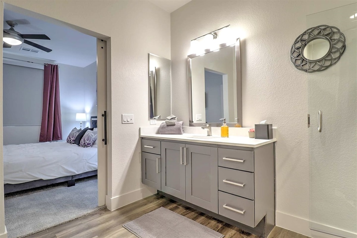 - Step into a modern ensuite bathroom with sleek gray cabinets and soft lighting   - Spacious layout connects directly to the cozy bedroom for added comfort   - Designed for ease with bright mirrors, clean finishes, and relaxing atmosphere
