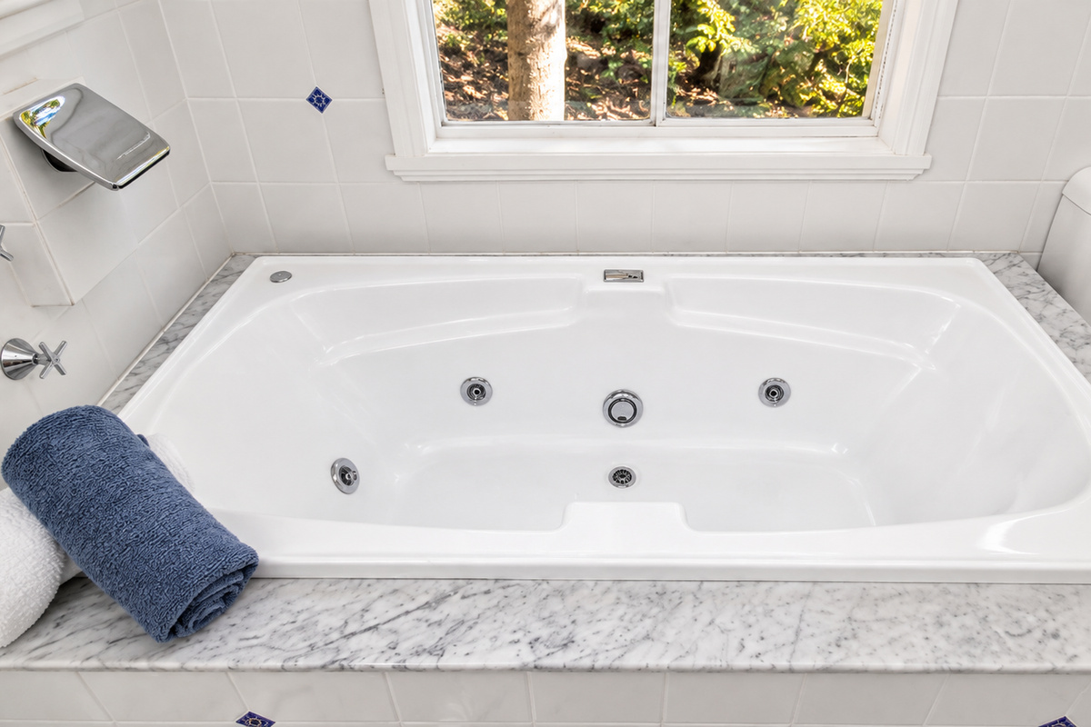 The bright bathroom features a spacious  bath surrounded by marble-look finishes and a peaceful window overlooking the greenery outside.