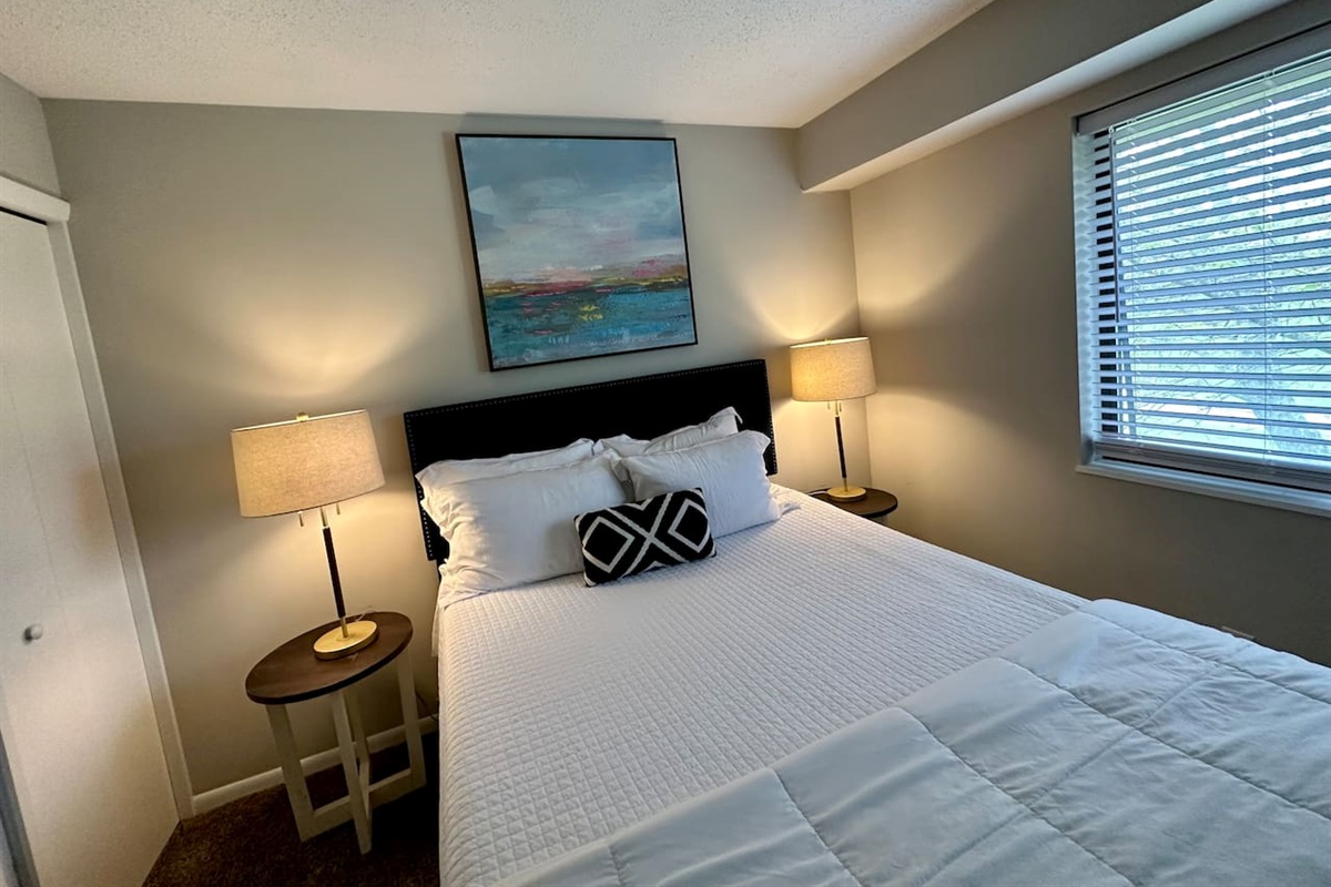 Unwind in this serene bedroom retreat featuring a comfortable queen-size bed, crisp white bedding, and warm accent lighting. A large window fills the room with natural light, while modern decor and blackout shades ensure a peaceful night's rest.