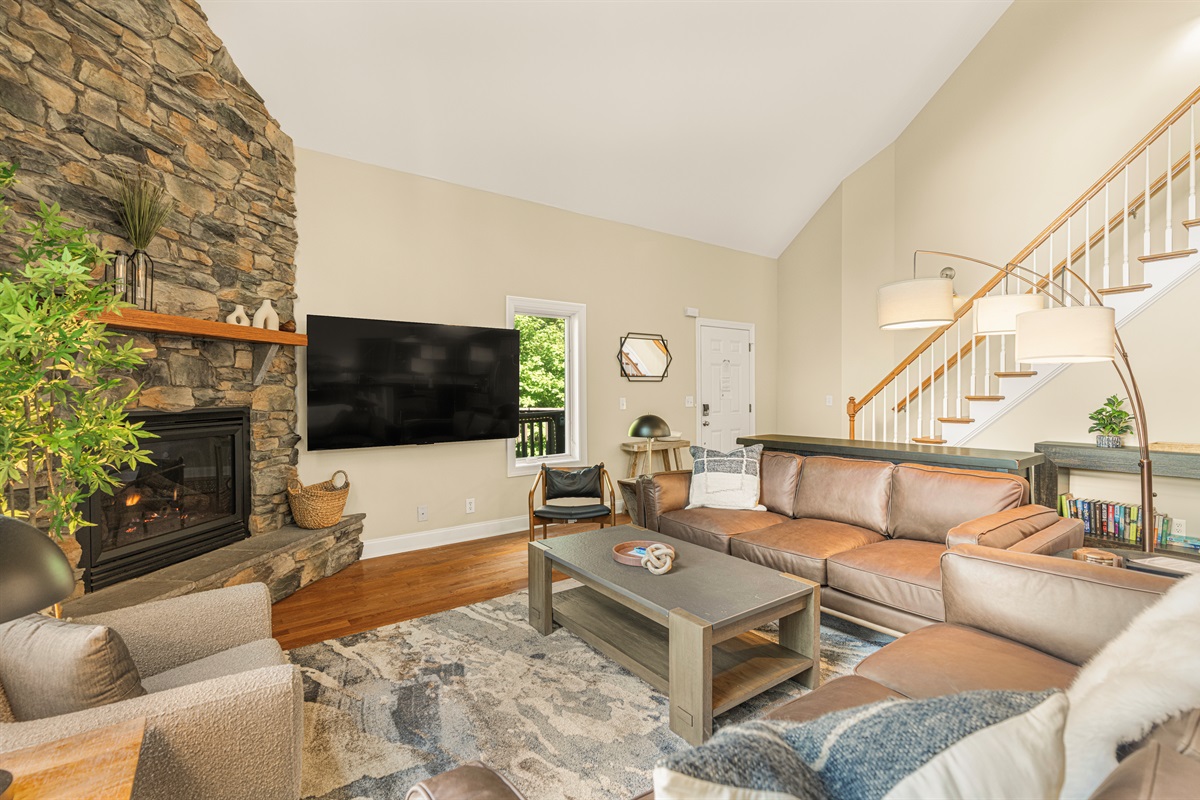Spacious vaulted living room with two oversized leather sofas, smart TV, stone gas fireplace, and natural light—perfect for cozy evenings or streaming your favorite shows in comfort
