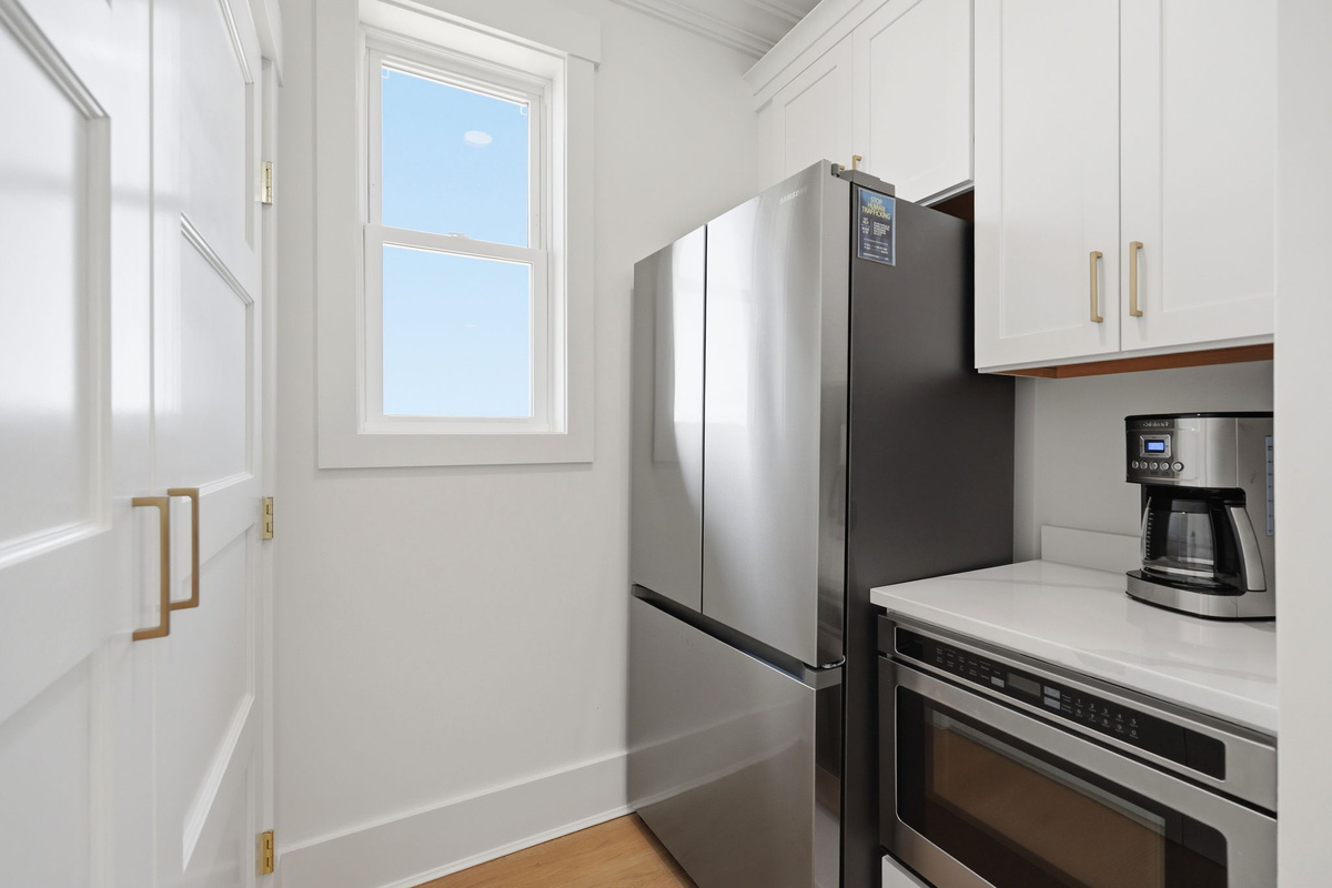 Compact secondary kitchen/utility area with stainless steel refrigerator, built-in oven, coffee station, and bright window-lit storage nook