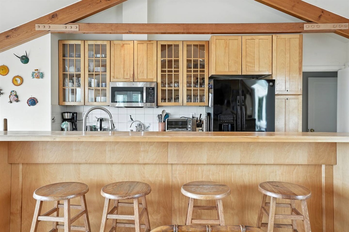 - Kitchen and breakfast bar combo with seating for four, perfect for casual dining  
- Features wood paneled cabinetry, black refrigerator, and open shelving style  
- Exposed beams and wall decor create a warm, welcoming coastal kitchen vibe