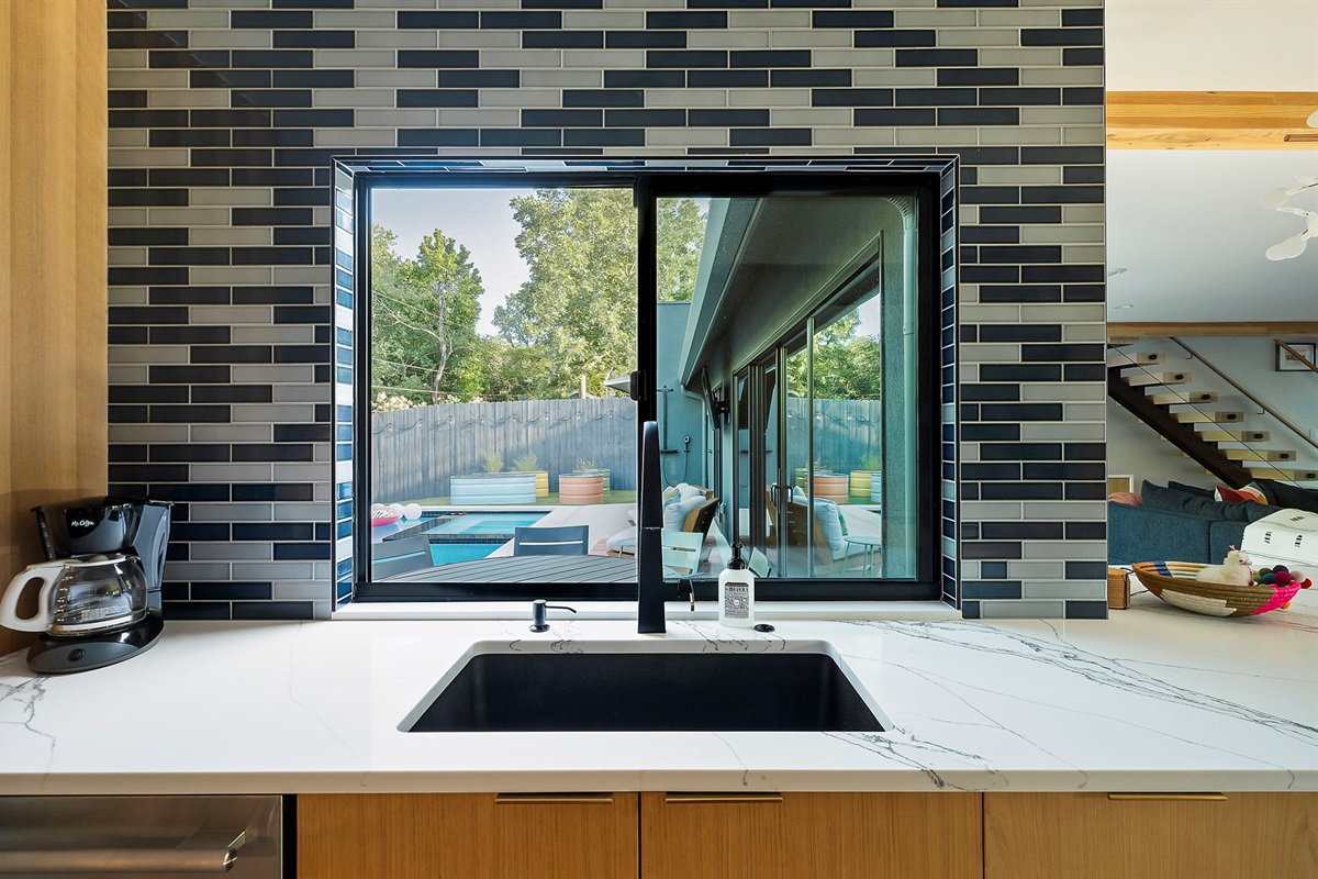 A modern kitchen featuring a black and gray subway tile backsplash, a large window overlooking a backyard with a swimming pool and a deck, and a deep sink with a contemporary black faucet set into a white marble-look countertop.
