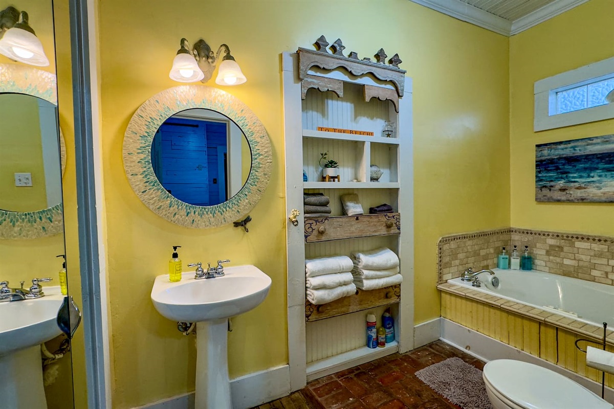 Charming bathroom featuring a tub/shower combo, open shelving with linens, and sunny, beach-inspired decor.
