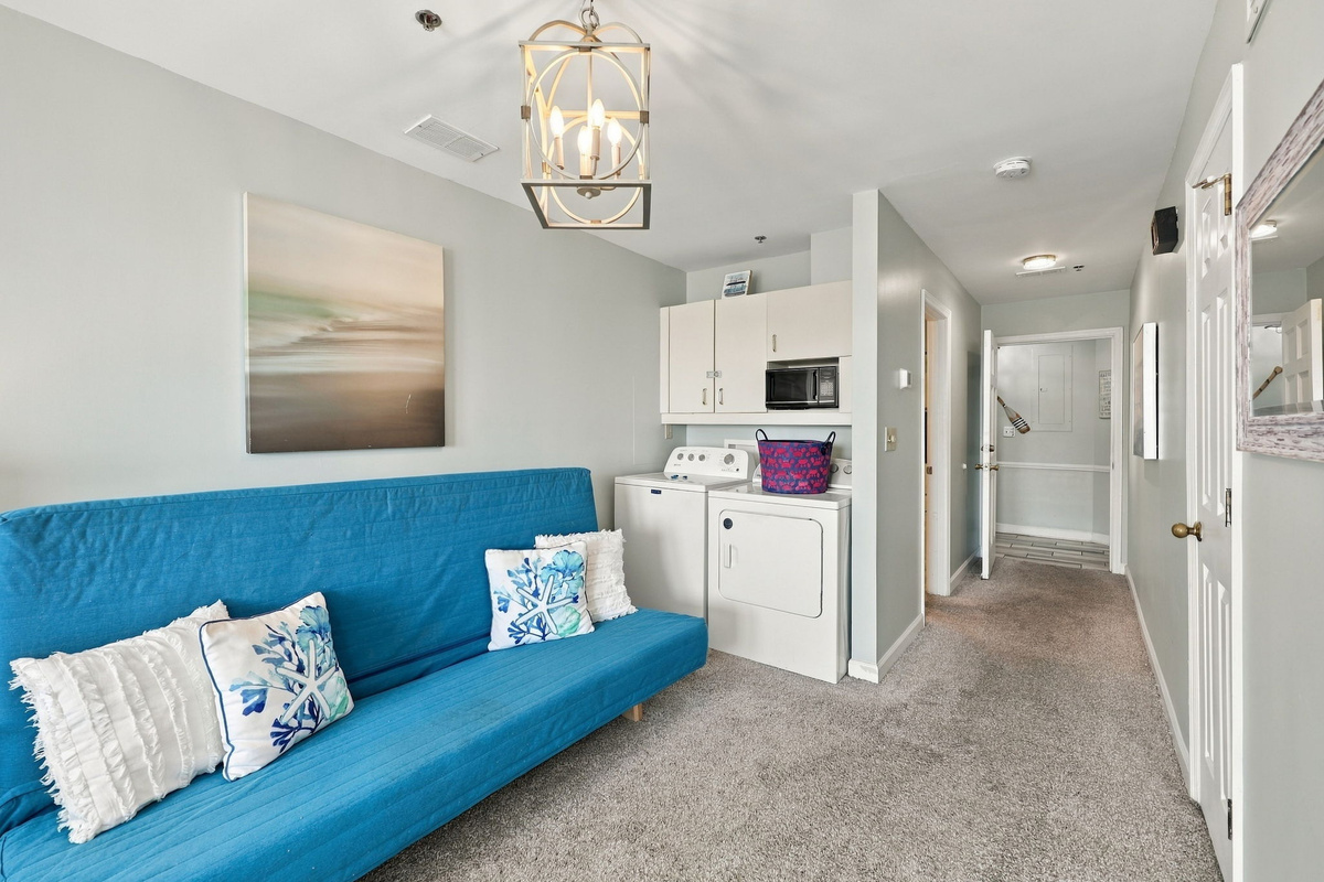 Versatile bedroom sitting area featuring a bright blue sofa bed, cage lantern light fixture, serene coastal wall art, and an integrated laundry nook with washer, dryer, and storage cabinets.   
