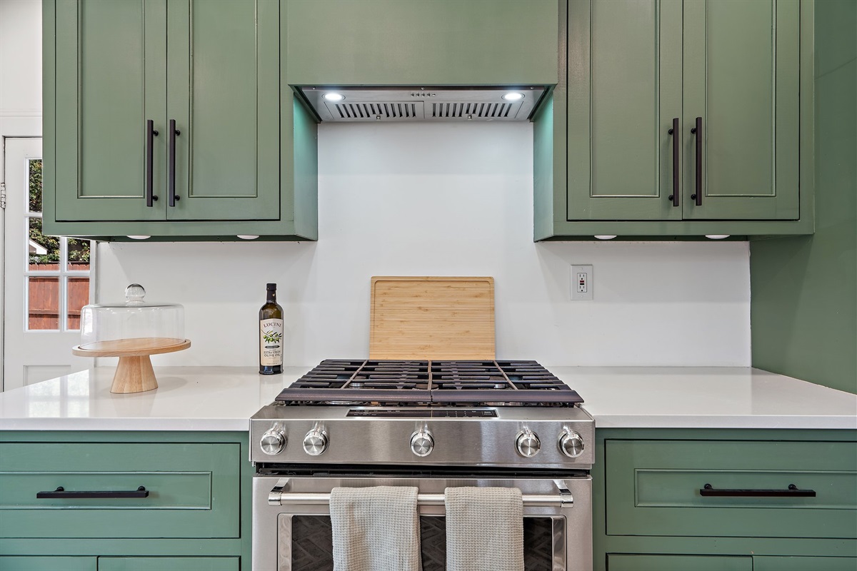 Kitchen gas range with range hood, sage green cabinetry, and butcher block accents. Full cookware, cooking basics, wine glasses, dishwasher, and Keurig coffee maker included. Main house, first floor.