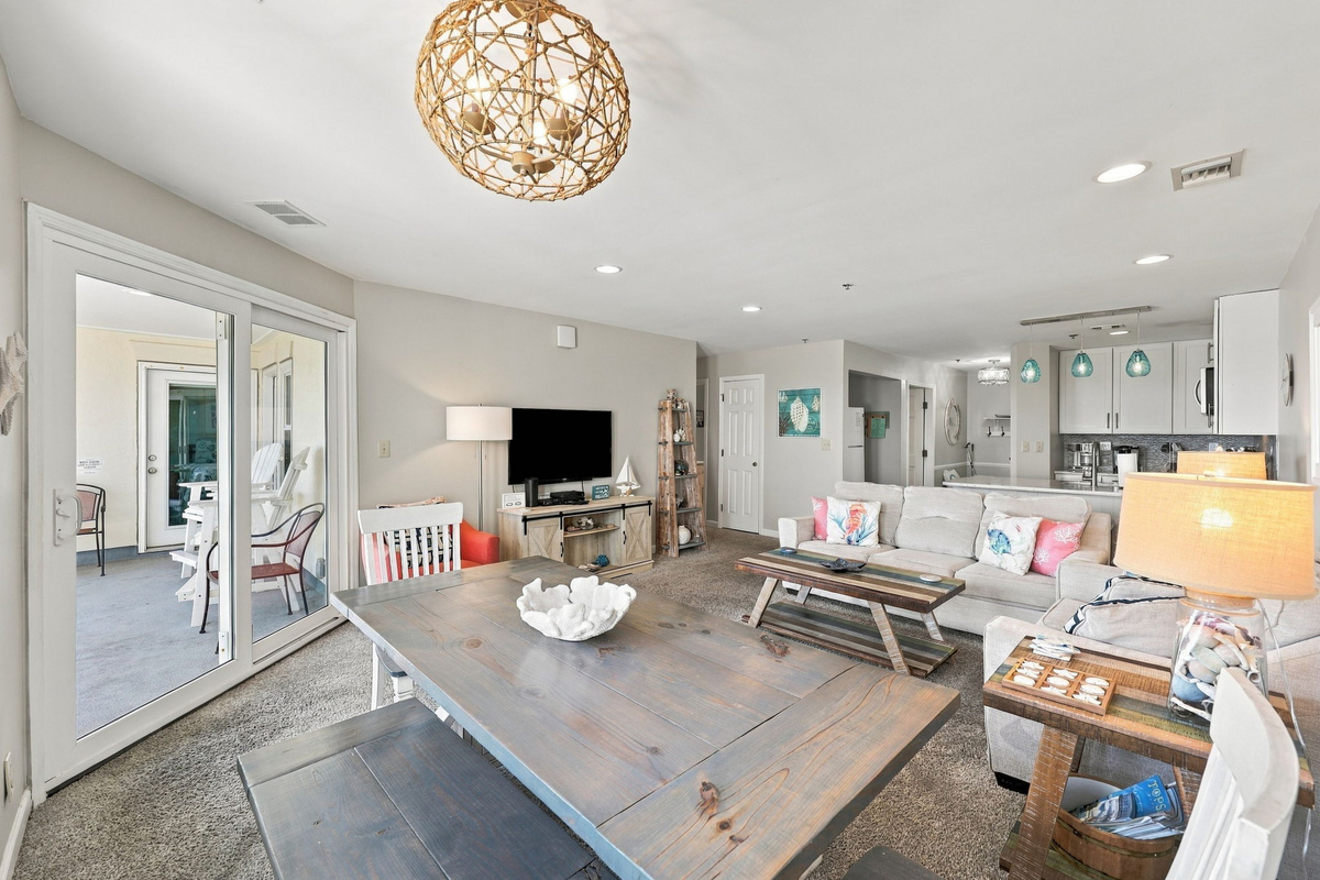 Sunlit dining and living space featuring a rustic wood table, woven chandelier, comfortable seating, and sliding glass doors opening to a covered patio.